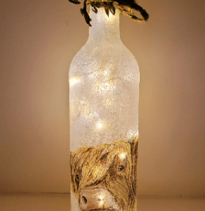 Standard wine bottle with Highland Cow detail on either side, jute twine with leaves around the neck, lit up with cork LED lights