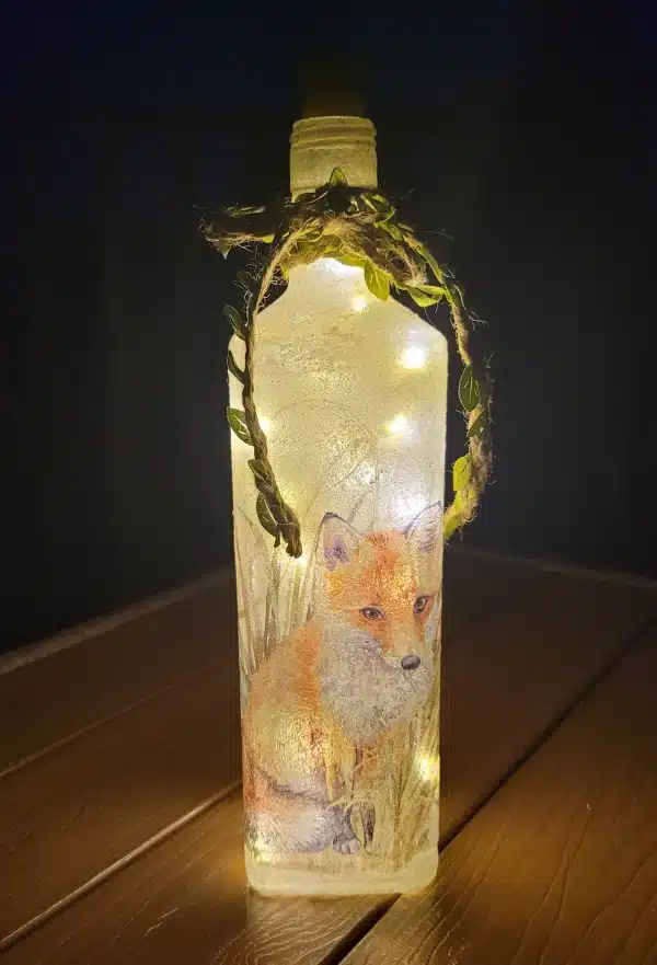 Lit up square gin bottle with fox detail and jute twine around the neck. Green leaves on the twine. cork LED lights